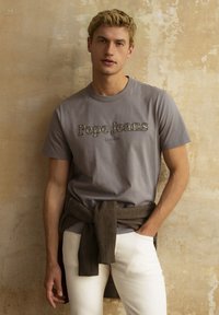 T-shirt en coton gris avec le logo "Pepe Jeans London", portée avec un pantalon blanc et un pull à côtes marron noué autour de la taille.