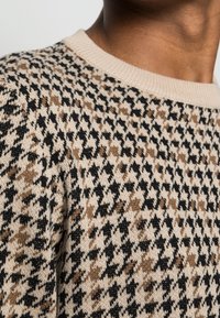 Sweter dzianinowy z cekinowym wzorem w odcieniach beżu, czerni i brązu. Okrągły dekolt oraz teksturowana tkanina o miękkim wykończeniu.
