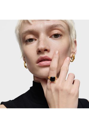 Anillo de oro con una piedra cuadrada negra, con acentos redondeados. El modelo lleva un suéter negro de cuello alto y pendientes de aro dorados, mostrando la colocación de la mano.