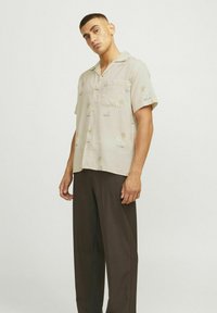 Kurzärmliges, hellbeiges Hemd mit floralem Muster und Tasche, kombiniert mit dunkelgrauen, lockeren Hosen, die einen entspannten Look schaffen.