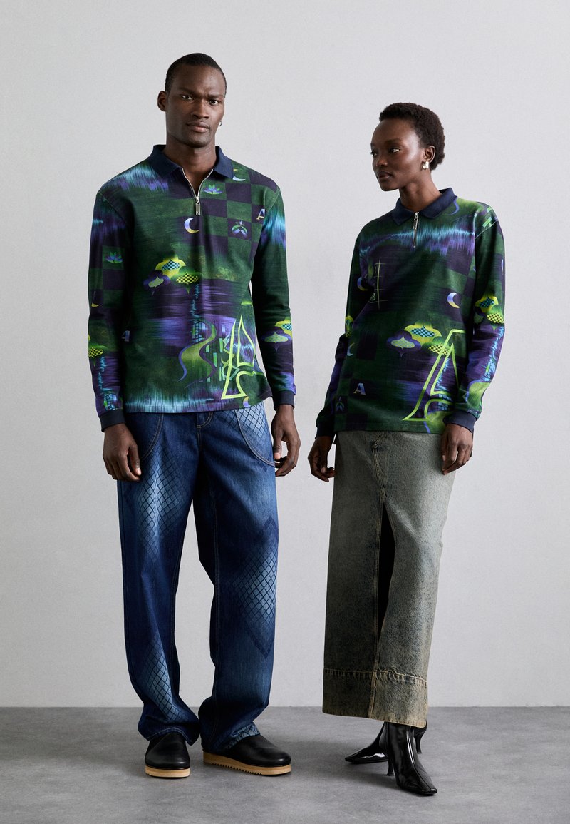 To personer bærer matchende mønstrede langærmede polo-shirts i nuancer af grøn og lilla, med grafiske designs. Manden parrer sin med denimbukser; kvinden har en nederdel på. Begge har forskellige fodtøj.