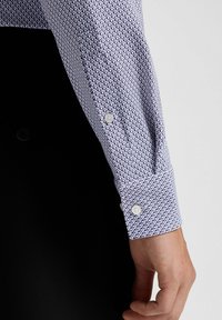 Langärmliges, tailliertes Hemd mit einem geometrischen Muster in Marineblau und Weiß. Sichtbare Knopfmanschetten, mit glatter Baumwollstruktur. Schwarzer Hintergrund.