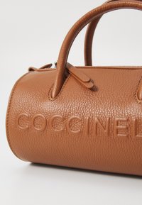 Braune Leder-Handtasche mit strukturierter Oberfläche, mit runden Griffen und geprägtem Branding auf der Oberfläche. Kompakte zylindrische Form.