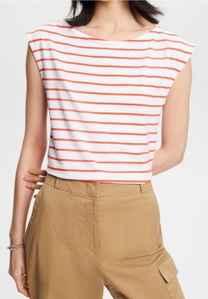Kobieta w białej bluzce bez rękawów z pomarańczowymi poziomymi paskami i wysokiej talii beżowych spodniach typu cargo, stojąca z jedną ręką za plecami.