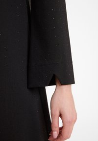 Zwarte jurk van een gestructureerde stof, met zilveren stipdetails. De mouw heeft een uitsnede bij de manchetten, waar een hand met manicured nagels te zien is.