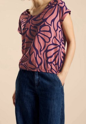 Femme portant un haut rose ample avec des motifs abstraits de feuilles violet foncé et un pantalon en jean bleu foncé, une main dans la poche.