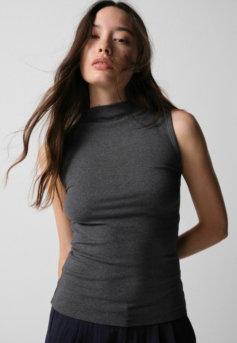 Young woman with long brown hair wearing a sleeveless dark gray top standing against a plain light background.