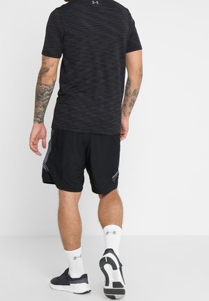 Mężczyzna w czarnej sportowej koszulce i krótkich spodenkach, białych skarpetkach i czarnych butach sportowych, stoi twarzą do białej ściany, z widocznymi tatuażami na ramionach.