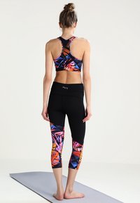 Kobieta stoi boso na szarej macie do jogi, ubrana w czarne legginsy 3/4 i kolorowy, wzorzysty stanik sportowy, zwrócona plecami w stosunku do jednolitego tła.