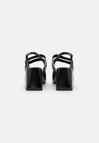 Sandalias de tacón de charol negro con tacón cuadrado, múltiples tiras en la parte trasera y detalles de hebilla ajustable en el tobillo.