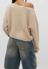 Beżowy sweter dzianinowy o luźnym kroju, teksturowanym ściągaczu i fasonie odsłaniającym ramiona, zestawiony z niebieskimi szortami denimowymi.
