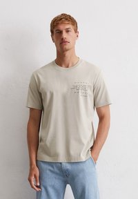 Light beige cotton t-shirt with short sleeves, crew neck, and text accents on the front. Paired with light blue denim jeans.