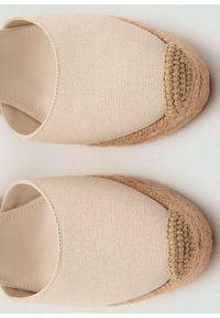 Des chaussures slip-on en toile beige avec un bout arrondi, présentant un accent en jute tissé autour de la base et un upper en tissu texturé.