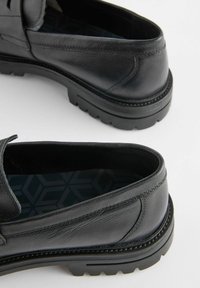 Sorte læder slip-on sko med en tekstureret overflade og gummisål, med en lav profil og en geometrisk mønstret sål.