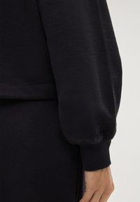 Felpa nera con una vestibilità leggermente ampia, polsini a costine e una texture liscia. Presenta un design semplice senza ulteriori motivi o accenti.