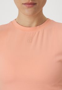 Top de manga corta de color rosa con nervaduras que presenta un sutil detalle de logotipo en relieve en el pecho y un cuello redondo. Textura de tela suave y elástica.