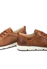 Zapatillas de cuero marrón con diseño perforado, suela beige, cordones marrones y detalle del logo en el lateral. Presentan una forma clásica y casual.