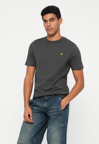 Homem a usar uma t-shirt cinza escura de mangas curtas com um pequeno logótipo de um pássaro amarelo e uns jeans azuis largos, em pé contra um fundo branco.