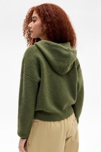 Sudadera con capucha verde hecha de un tejido texturizado. Presenta mangas y dobladillo acanalados, con un corte holgado y un diseño informal, combinada con pantalones claros.