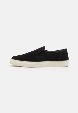 Chaussures noires en daim à enfiler, avec un bout rond, une semelle en caoutchouc crème et sans lacets ni fermetures visibles. Texture lisse, design minimal.