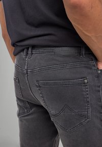 Dark gray denim jeans featuring a leather logo patch, prominent stitched pockets, and flat rivets. The fabric has a slightly worn texture.