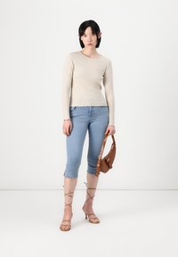 Beige ribbet lange ermers topp med bølget kant, kombinert med lyseblå denim capri bukser og beige høyhælte sandaler. Brun skulderveske i hånden.