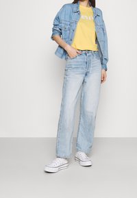 Světle modrá džínová bunda, žluté tričko s logem, světle modré džíny a bílé tenisky. Neformální outfit s volným střihem.
