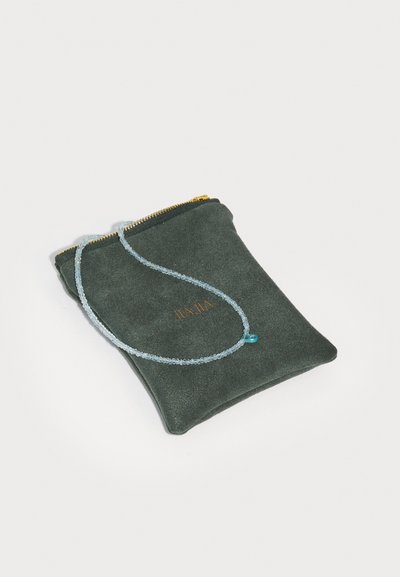 Žalias suede maišelis su auksiniu užtrauktuku, laikantis subtilią mėlyną brangakmenių vėrinį, kuriame yra mažas širdelės formos pakabukas.