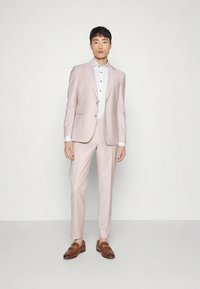 Costum roz deschis dintr-un material fin, cu croială mulată. Asortat cu o cămașă albă cu nasturi negri și pantofi maro tip loafer.