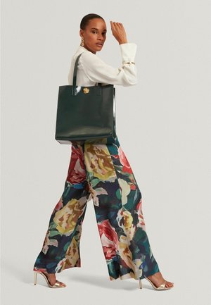 Bolsa tote verde brillante con dos asas alargadas y un detalle en dorado. La modelo lleva una blusa blanca y pantalones anchos de flores coloridas.