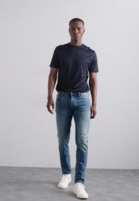 Fekete, rövid ujjú póló, kék, slim-fit farmer és fehér sneaker. Semleges háttér előtt állva mutatja be a laza öltözetet.