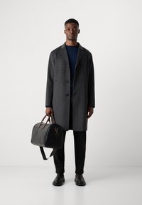 Donkergrijze wollen jas met een gepunt kraag, knoopsluiting en twee voorzakken. Zwart leren duffeltas met bruine leren accenten. Zwarte broek en schoenen.