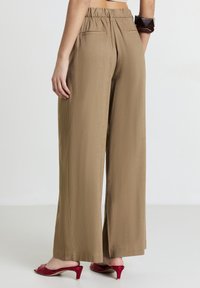 Donna che indossa pantaloni beige a vita alta e gamba larga abbinati a mules con tacco rossi, con braccialetti in vista sul polso.