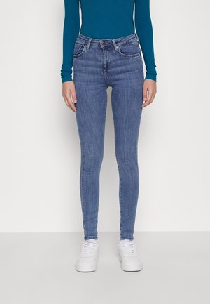 Woman wearing blue skinny jeans, teal long-sleeve top, and white sneakers, standing against a plain light background.