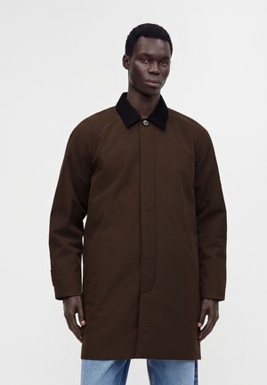 Homme portant un manteau marron foncé avec un col noir sur un jean bleu clair sur un fond blanc uni.