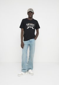 Crna grafička majica s natpisom "ARCHIVE", uparena s širokim trapericama svijetloplave boje i bijelim tenisicama s sivim detaljima. Sivi šešir upotpunjuje odjevnu kombinaciju.