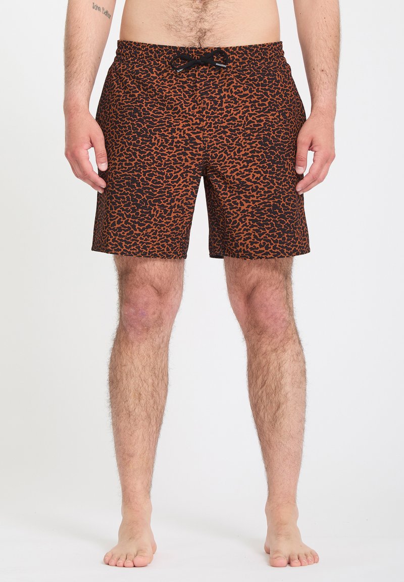 Homme portant un short de bain marron et noir à motifs avec un cordon noir, debout pieds nus devant un fond uni.