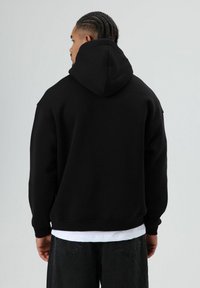 Zwarte hoodie met een comfortabele pasvorm, geribde boorden aan de mouwen en zoom. Zachte textuur zonder zichtbare patronen of logo's. Draagt over een wit shirt.