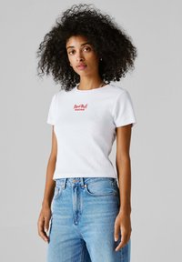 Kvinne med krøllete hår iført en hvit Red Bull Racing t-skjorte og blå jeans, stående mot en ensfarget grå bakgrunn.