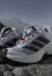 Λευκά και μαύρα παπούτσια Adidas Gore-Tex για τρέξιμο σε μονοπάτια, τοποθετημένα σε βραχώδη επιφάνεια με ορεινό τοπίο σε απαλό φόντο.