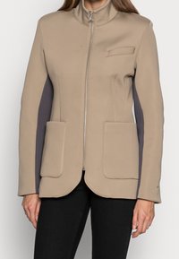 Chaqueta beige con cremallera y cuello alto, dos bolsillos tipo parche en la parte frontal y paneles laterales en gris oscuro, combinada con pantalones negros.