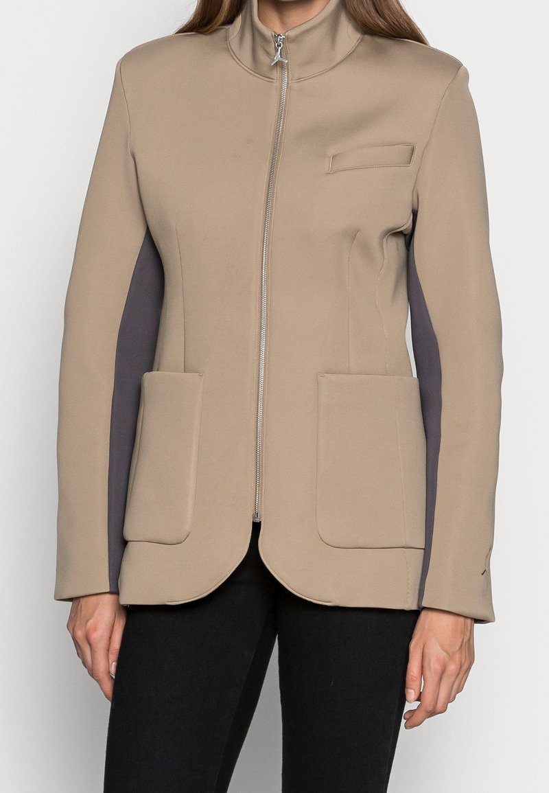 Chaqueta beige con cremallera y cuello alto, dos bolsillos tipo parche en la parte frontal y paneles laterales en gris oscuro, combinada con pantalones negros.