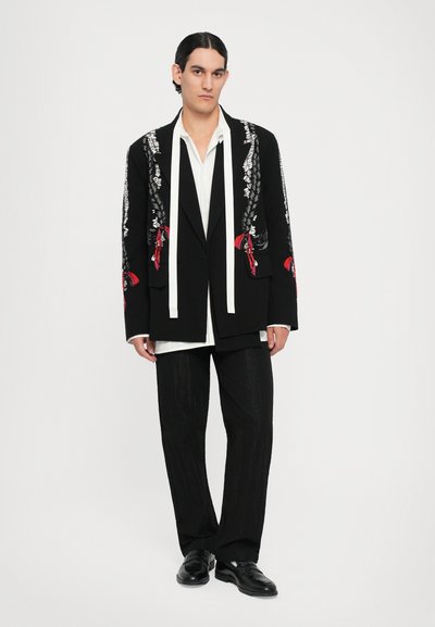 Schwarzer taillierter Blazer mit aufwändiger weißer und roter Blumenstickerei, kombiniert mit einem weißen Hemd und texturierten schwarzen Hosen, abgerundet mit schwarzen Schuhen.