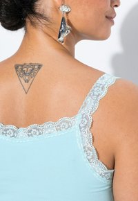 Ljusblå ärmössad med spetskant på axelbanden, som visar ett fint mönster. Närbild av triangulär tatuering på ryggen och iögonfallande örhängen.