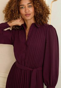 Burgundin pleated dress with a button-up front, collar, and tie waist. Long, puffed sleeves with textured fabric detail.