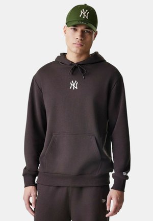 Dunkelbrauner Hoodie mit Fronttasche, Kordelzug und weißem Logo. Kombiniert mit einer grünen Kappe mit weißem Emblem. Lässige Sportbekleidung.