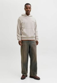 Hele beež kapuutsiga dressipluus esitasku ja paelaga; kantud koos lahtiste, tuhmide oliivroheliste teksade ja tumepunaste kingadega.