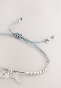 Verstellbares hellgraues Kordelarmband mit silbernen Perlen, kleinem Anhänger und zwei schlüsselartigen Anhängern auf hellem Hintergrund.