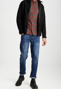 Czarna zamszowa kurtka, czerwona i szara koszula w kratę, niebieskie dżinsy z podwiniętymi nogawkami i czarne botki za kostkę. Skupienie na warstwowym, swobodnym wyglądzie.