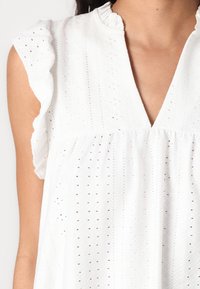 Blusa bianca con maniche corte a volant. Presenta un tessuto testurizzato con motivi in occhio di pernice e una scollatura a V. Silhouette fluida.